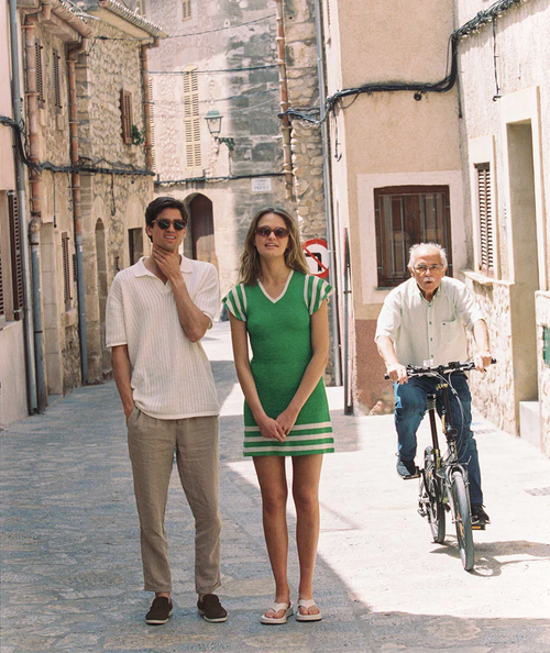A couple poses on a cobblestone street, with an elderly man riding a bicycle behind them, flanked by stone buildings and architectural details.