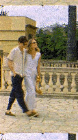 A couple walks along a stone path next to a balustrade on a sunny day, with buildings and green trees in the soft-focus background.