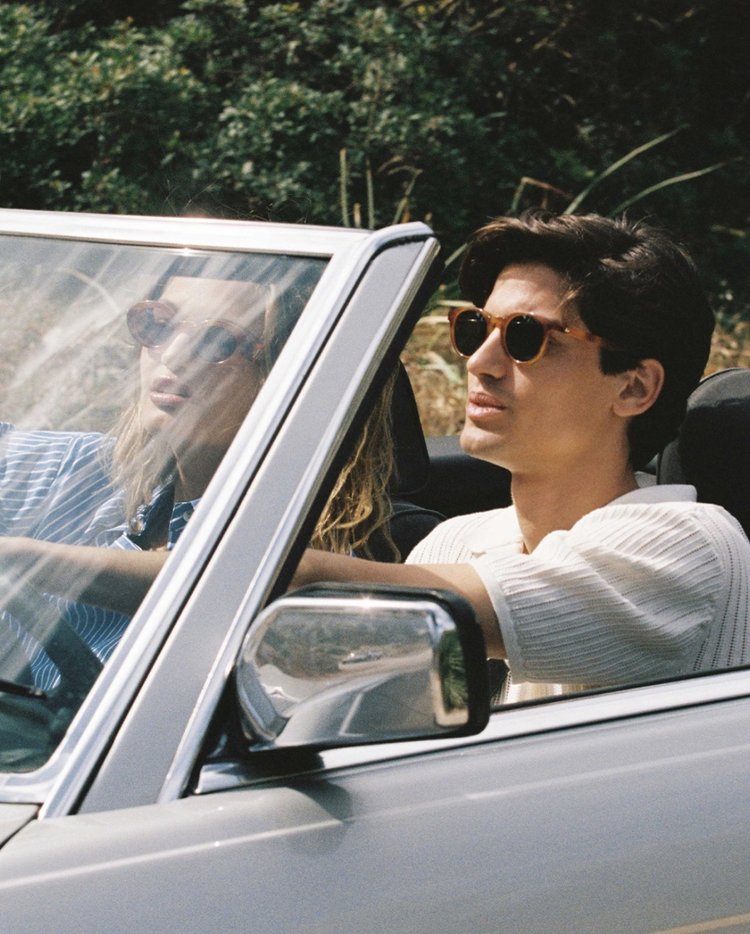 Man and woman wearing sunglasses driving a convertible car in a sunny outdoor setting for Le Specs An Ode to Summer campaign.