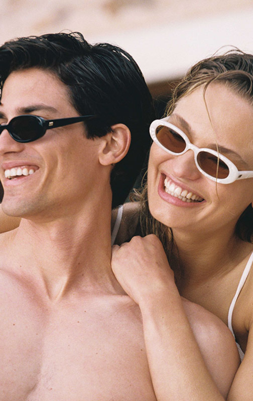Close-up portrait of a smiling man and woman wearing sunglasses outdoors in bright sunlight