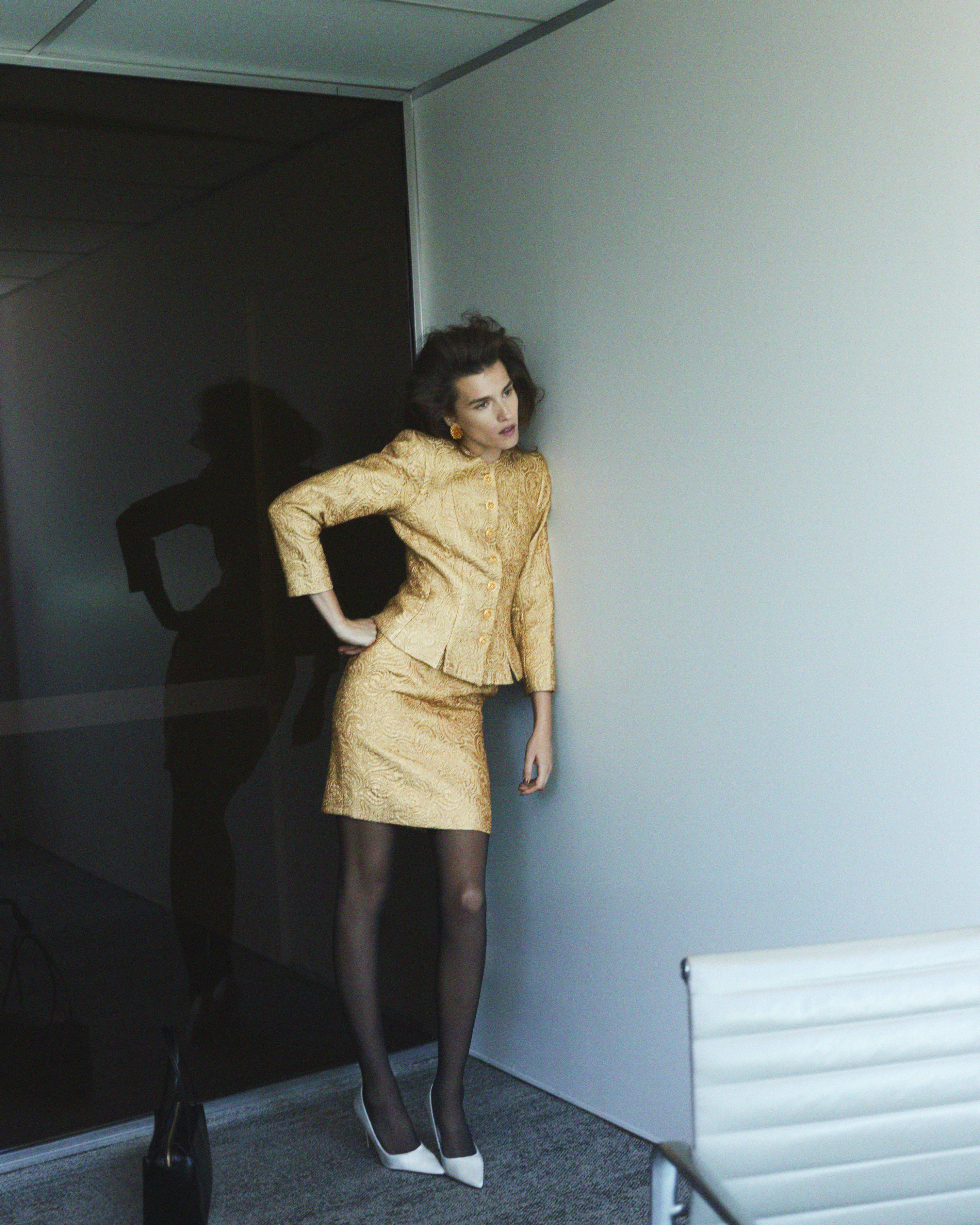 An adult woman in a gold skirt suit leans against a wall in an office interior. She wears black tights and white heels and has an introspective gaze.