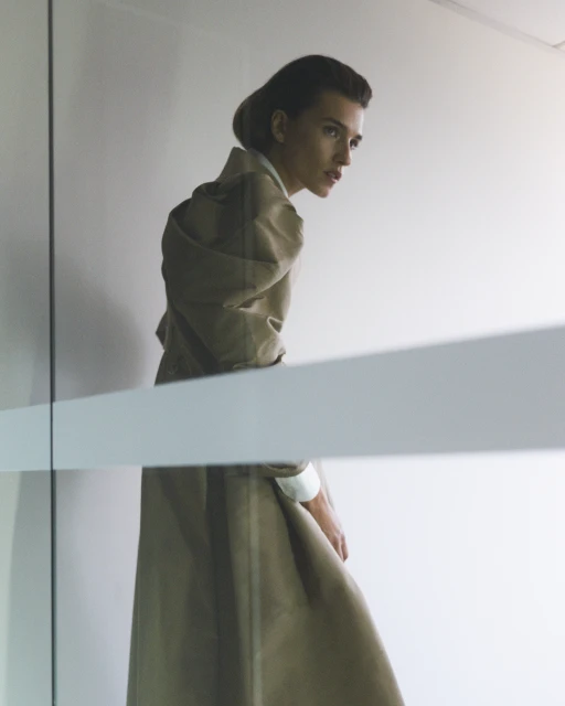 A woman in a long tan coat stands in front of a bright white wall partially obscured by frosted glass, looking away from the camera.