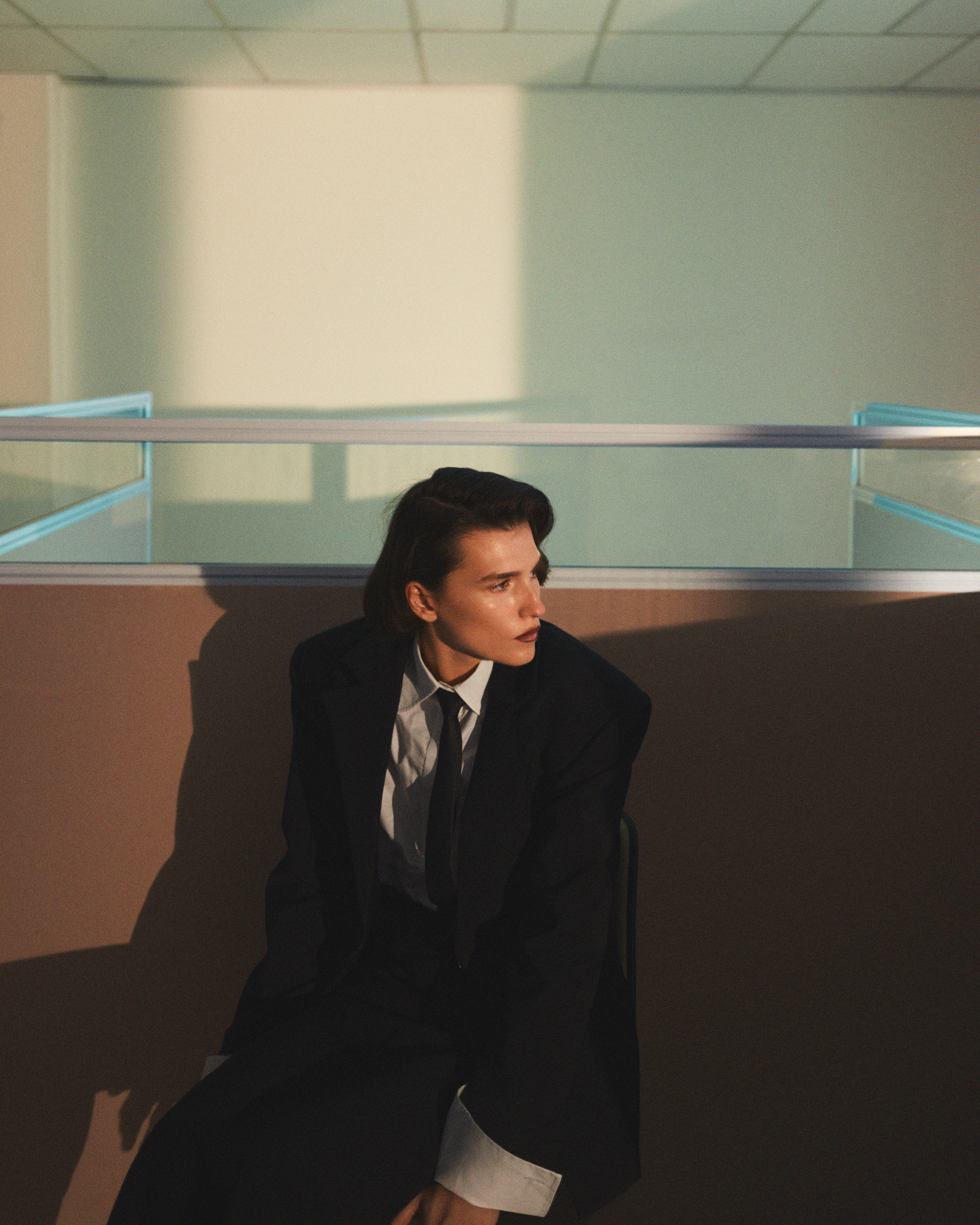 Portrait of a person with short hair in a dark suit, white shirt, and black tie, sitting indoors with sunlight and shadows.