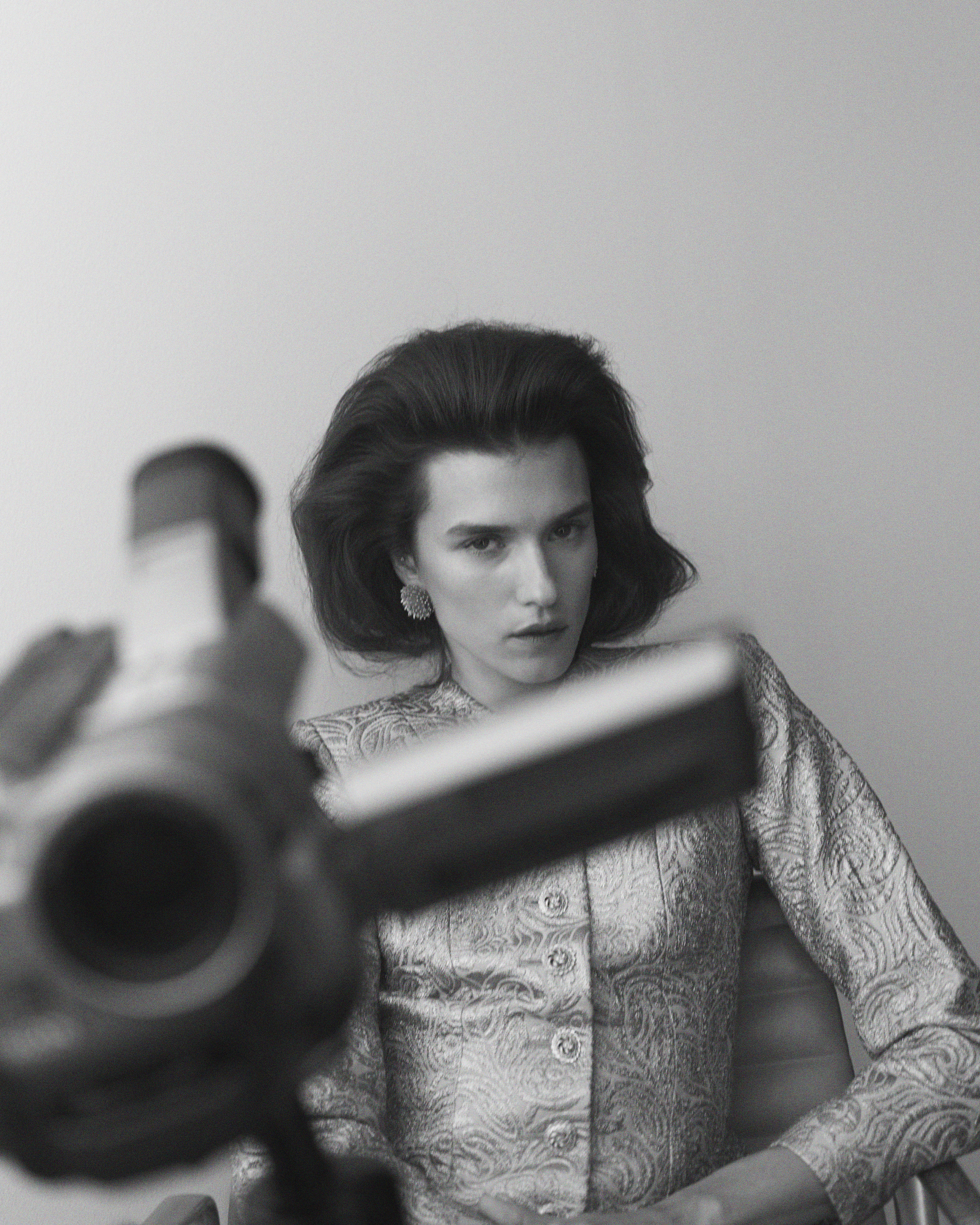 Monochrome portrait of a woman with short dark hair and pale skin, wearing patterned jacket and earrings, blurred camera in foreground.