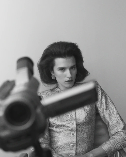 Monochrome portrait of a woman with short dark hair and pale skin, wearing patterned jacket and earrings, blurred camera in foreground.