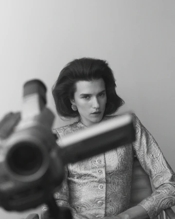 Monochrome portrait of a woman with short dark hair and pale skin, wearing patterned jacket and earrings, blurred camera in foreground.
