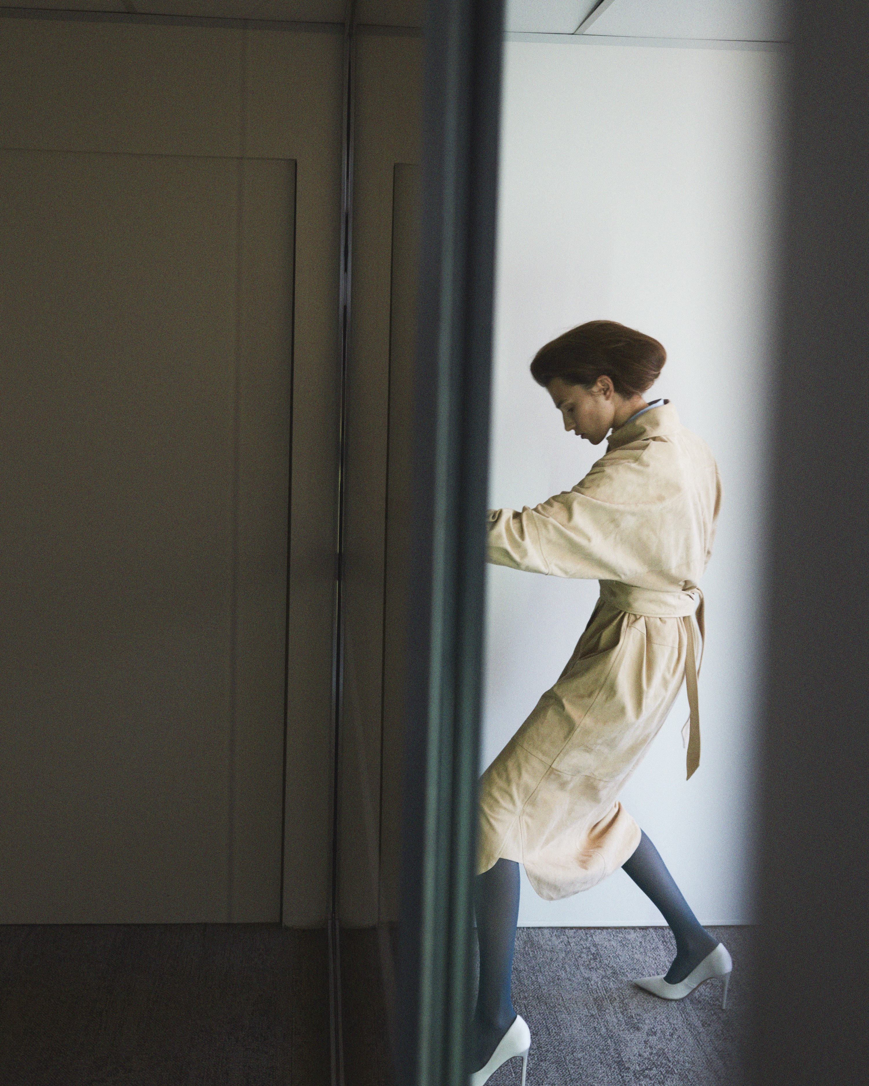 Model wearing beige trench coat and white heels standing indoors near dark reflective glass partition.