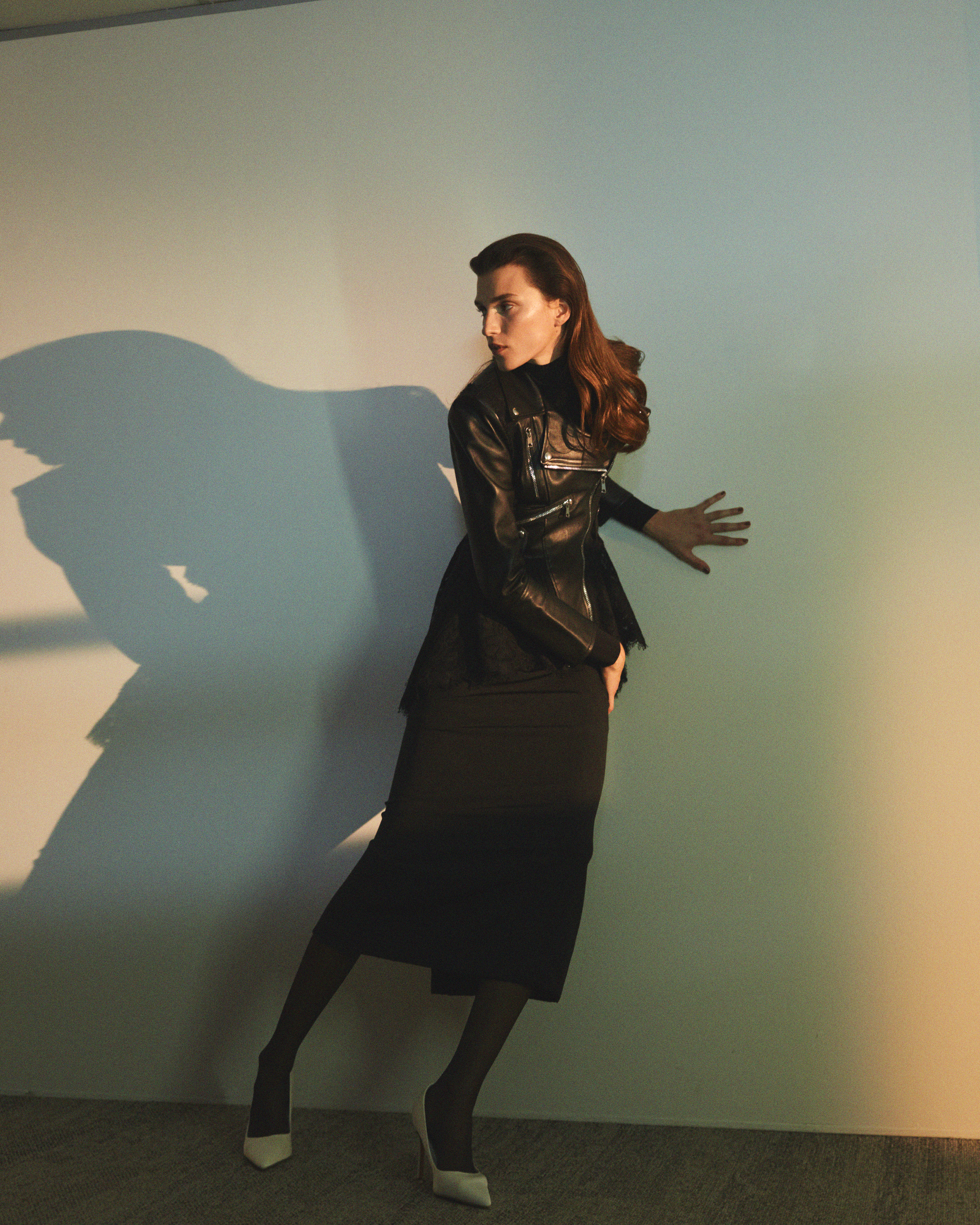 Woman in a black leather jacket and dark skirt posing against a wall with a prominent shadow for Numero Netherlands.