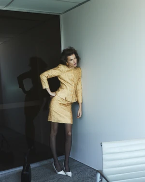 Model wearing a gold textured jacket and skirt leaning against an office wall in a fashion portrait for Numero Netherlands.