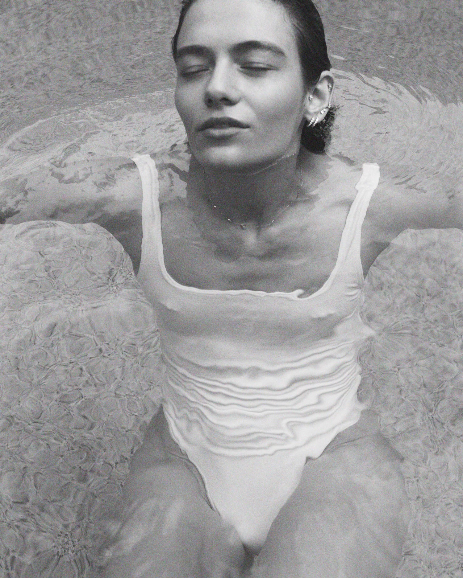 Monochrome portrait of a woman in a white swimsuit in water, eyes closed, silver ear cuff, serene mood, medium shot.