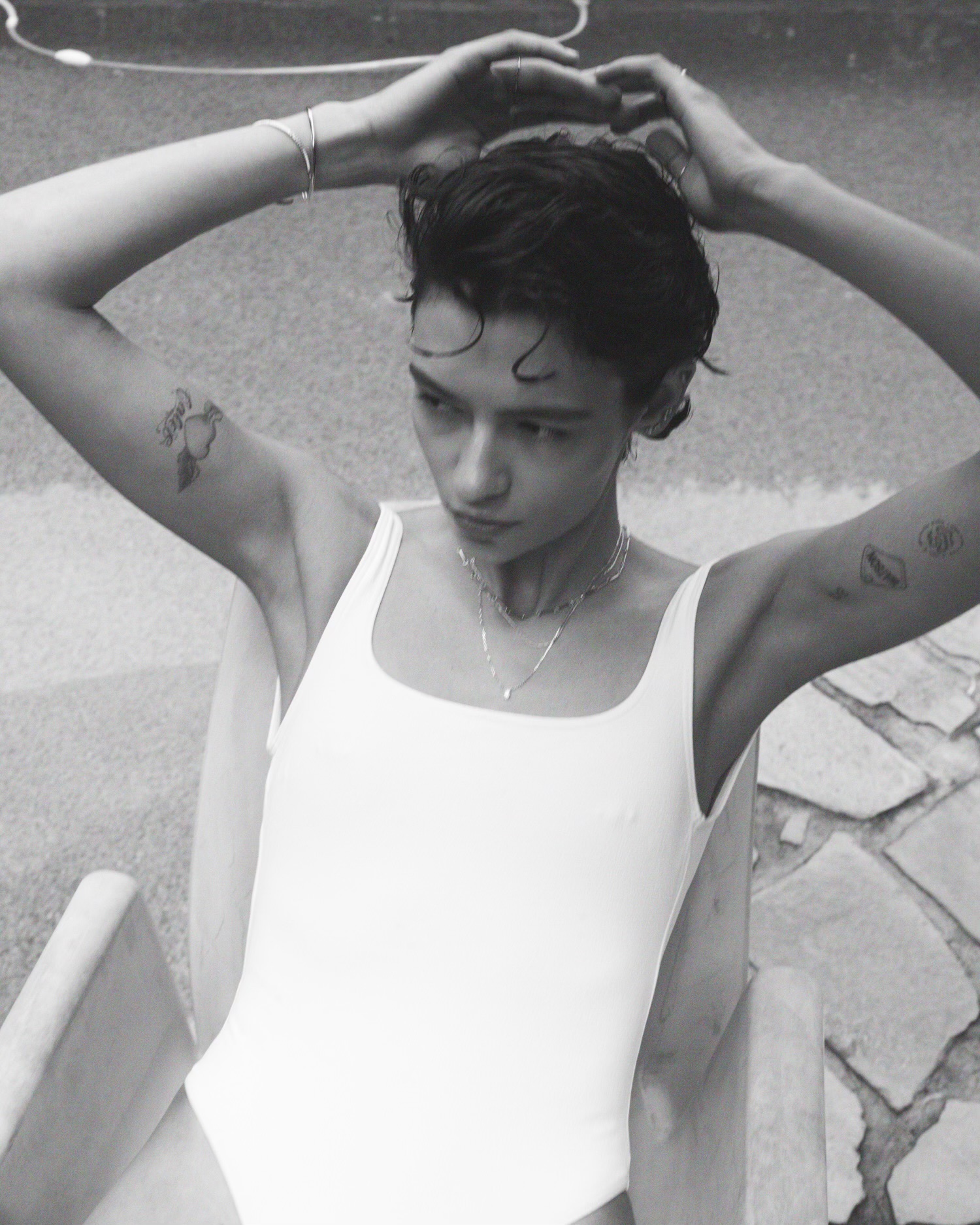 Black and white portrait of a woman wearing a white swimsuit and delicate layered necklaces, holding her hands behind her head.