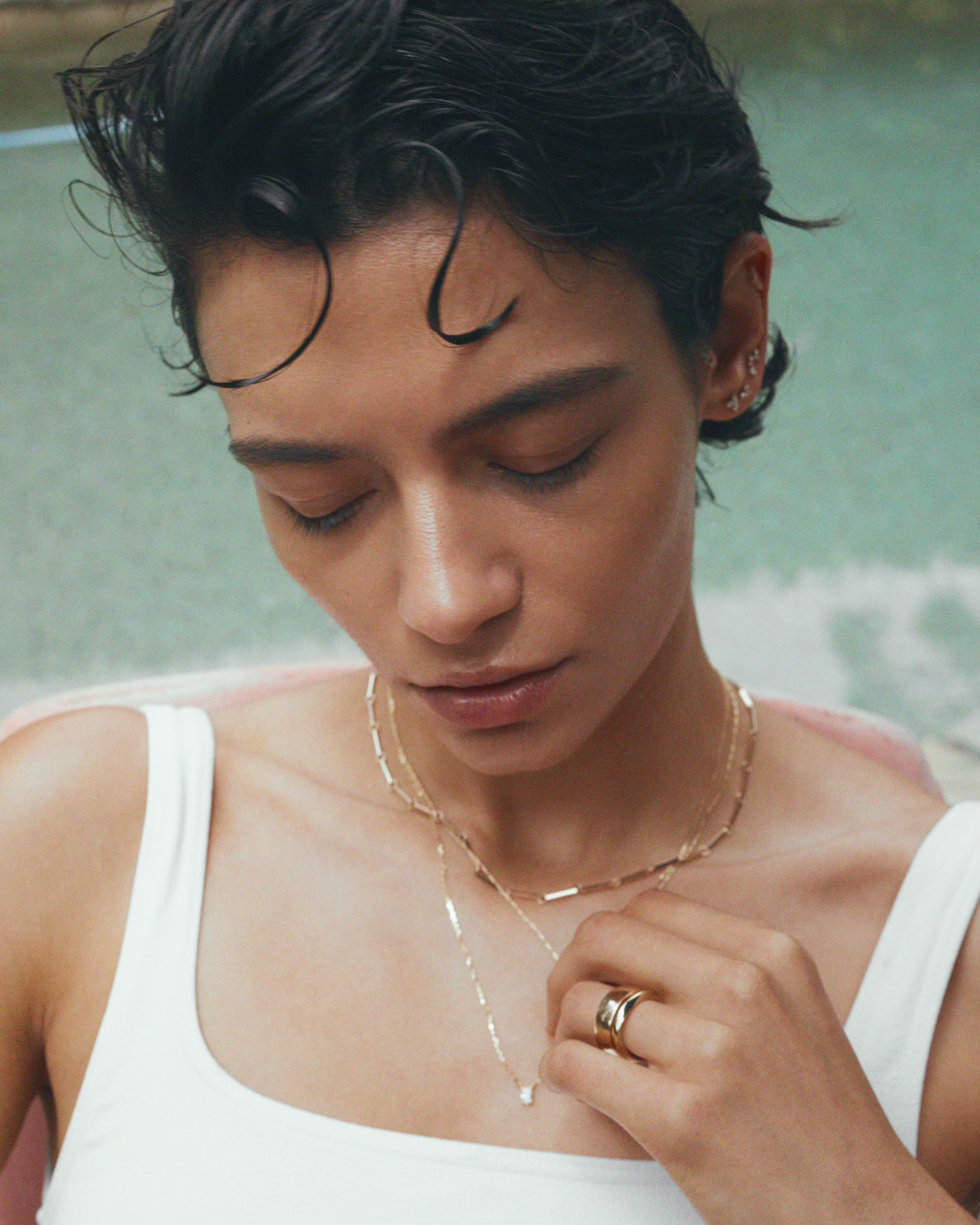 Close-up portrait of a woman wearing delicate gold necklaces and rings, featuring wet-look dark hair and a white tank top against a soft water background.