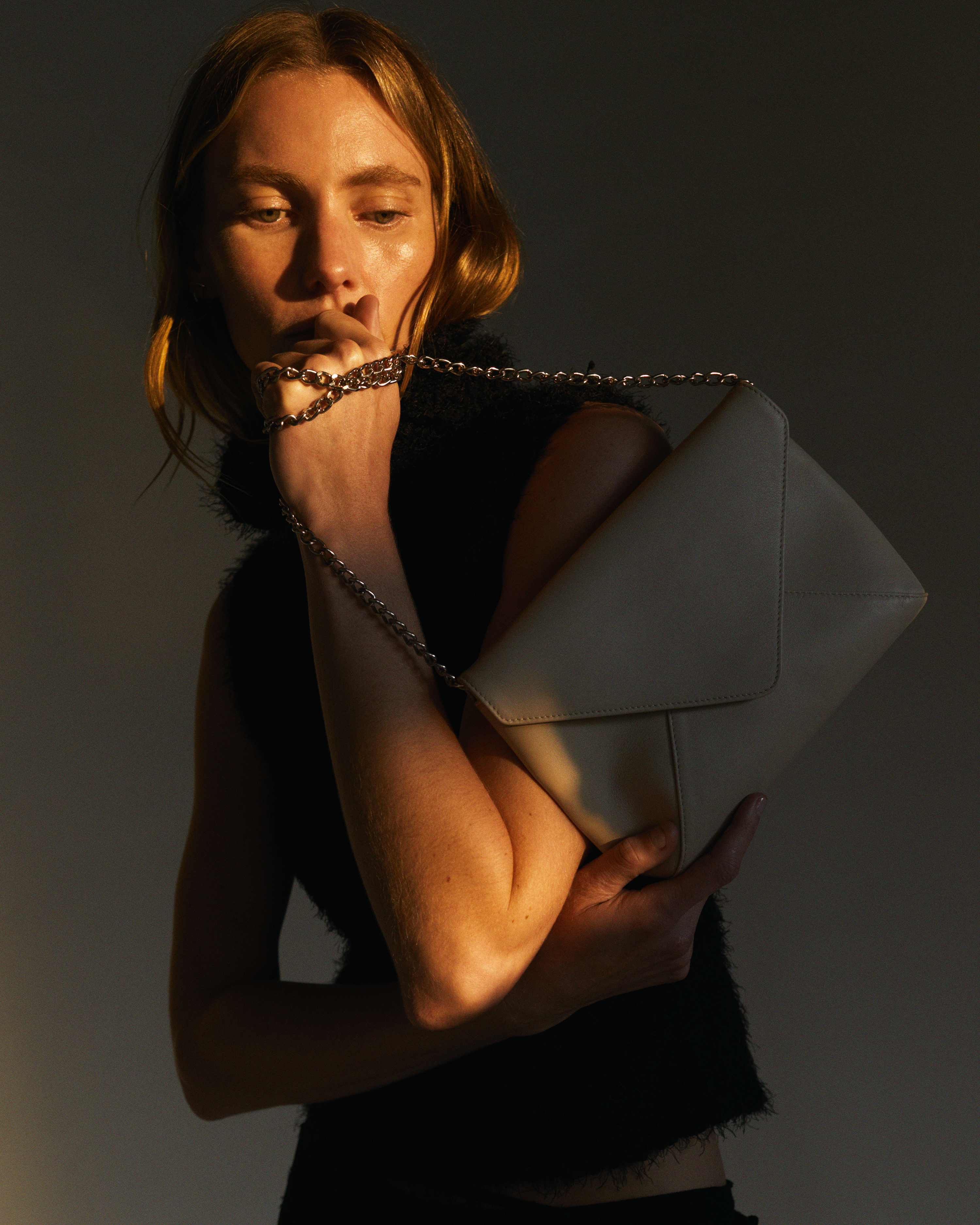 Close-up of a fair-skinned woman holding a beige handbag with a chain strap, warm lighting, and a dark, blurred background.