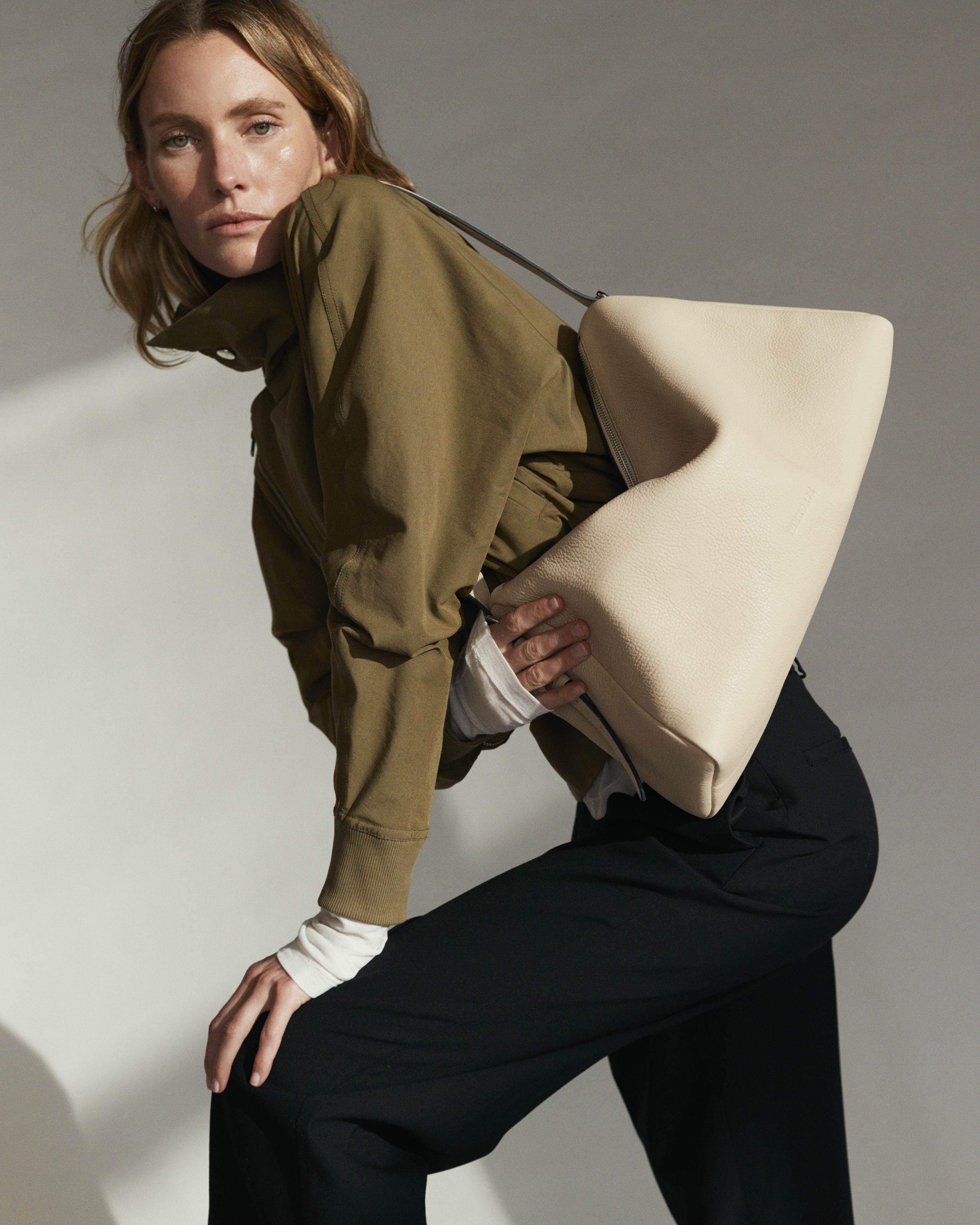 A woman poses in a light gray studio with a light beige hobo bag over her shoulder, wearing a khaki jacket and black pants.