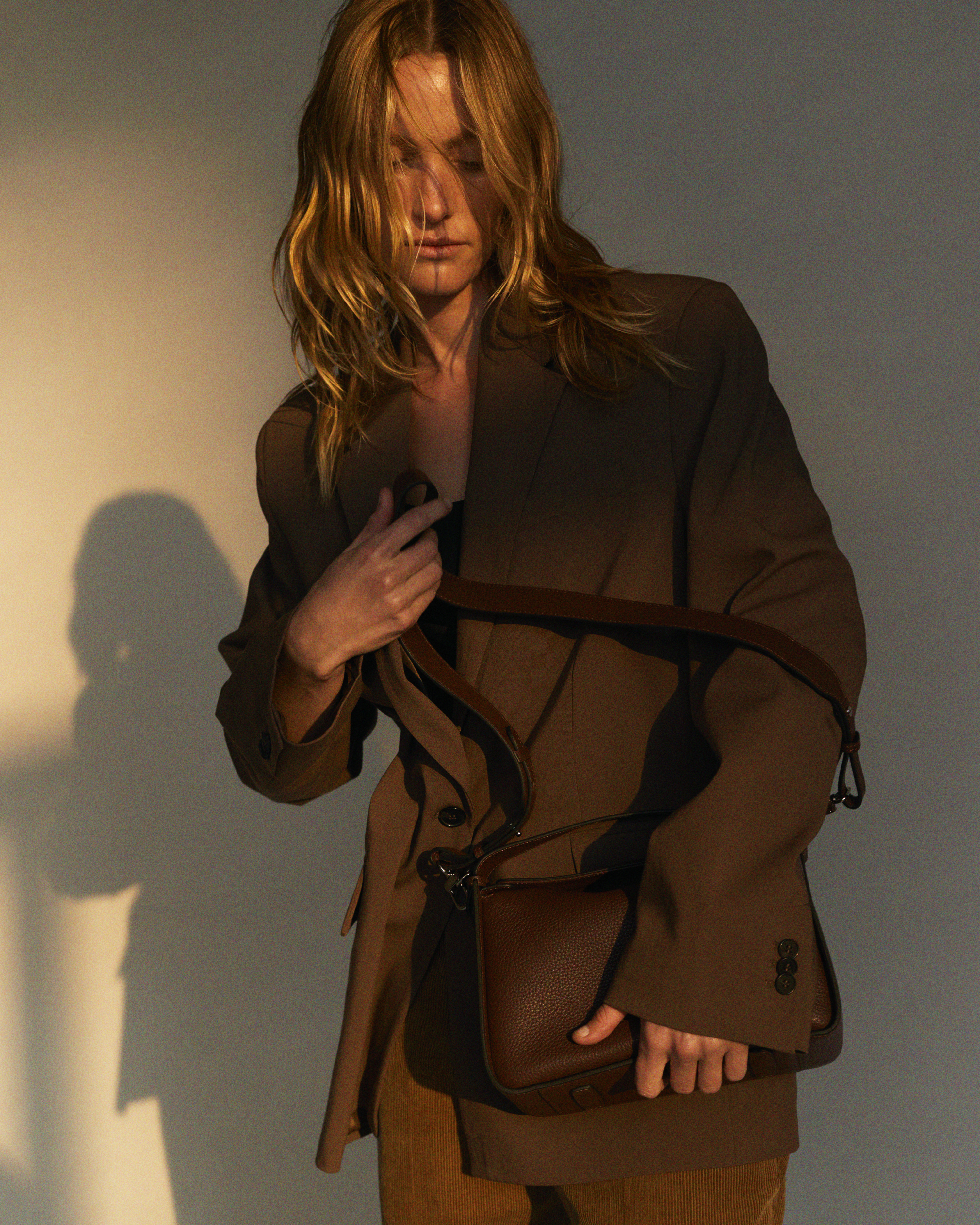 Woman in a brown blazer and brown pants holds a brown leather purse with a shoulder strap in a studio setting with warm light.