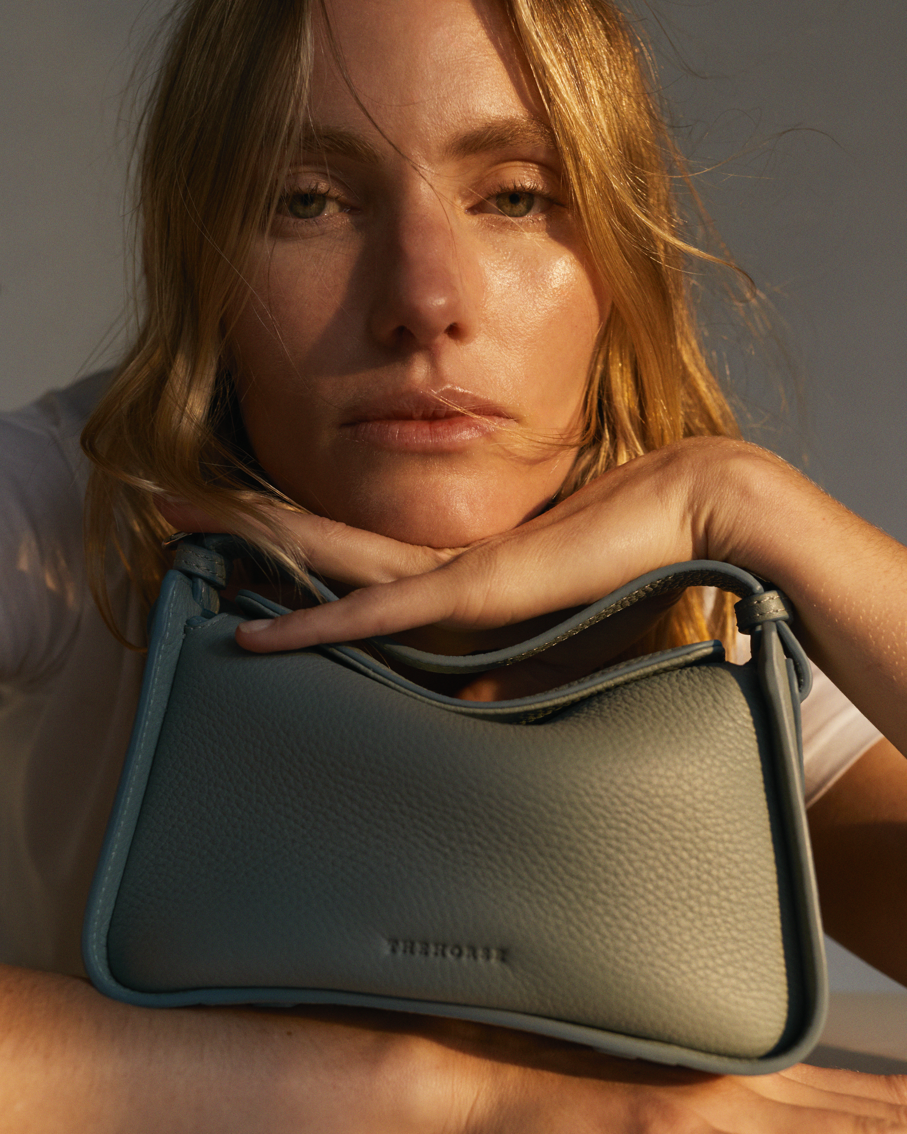 Close-up studio shot of a blonde adult female with light skin, resting her chin on a small, light blue leather handbag.