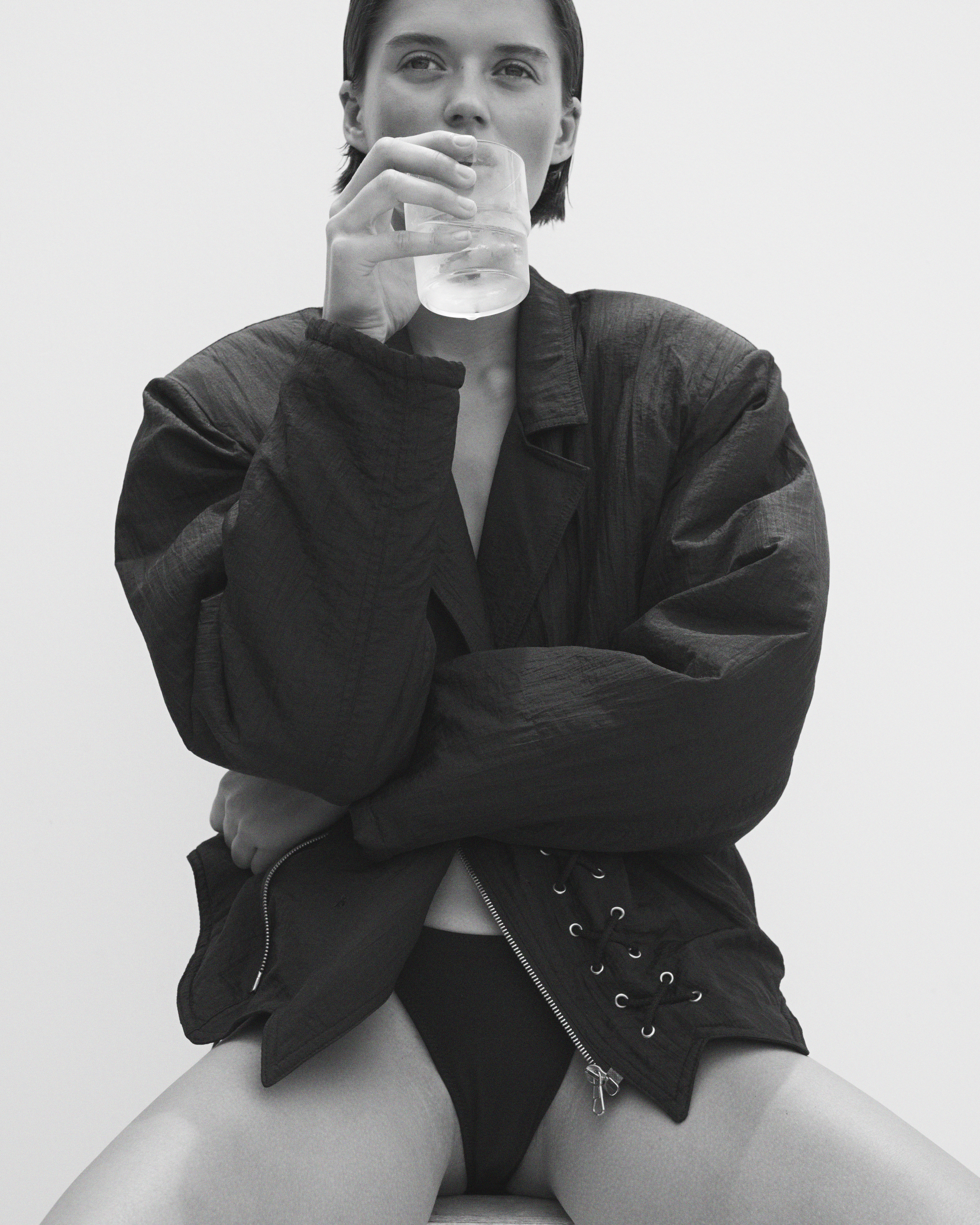 Black and white studio shot of a young adult woman in black jacket and underwear drinking water from a clear glass in a studio.