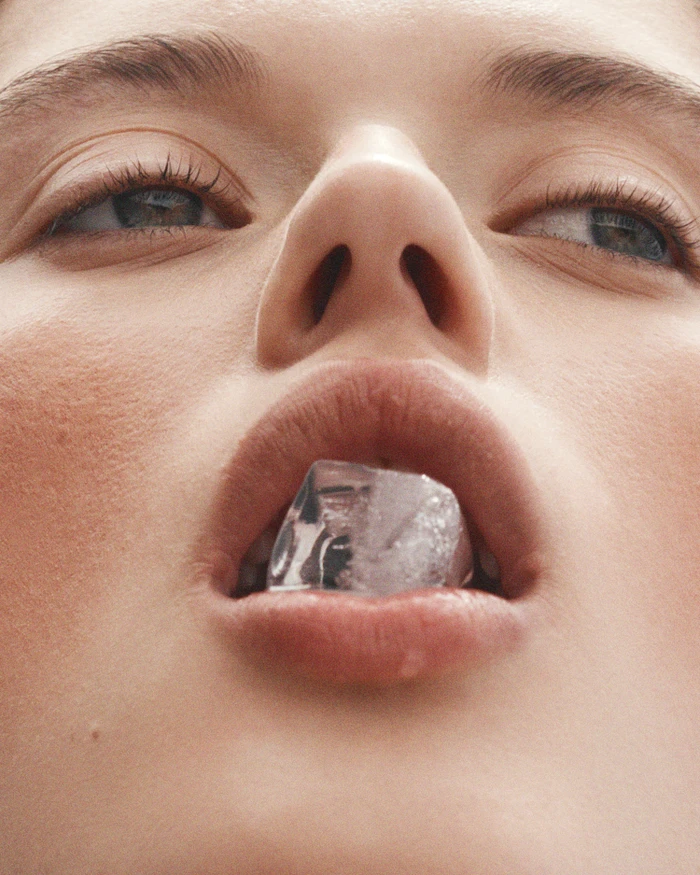 Extreme close-up portrait of a woman holding a single ice cube between her lips shot from below by photographer Tim Ashton.
