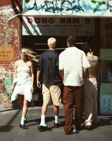 Group of four young adults in casual clothing standing on a city street in front of Dong Nam A Supermarket; brick building and sidewalk.