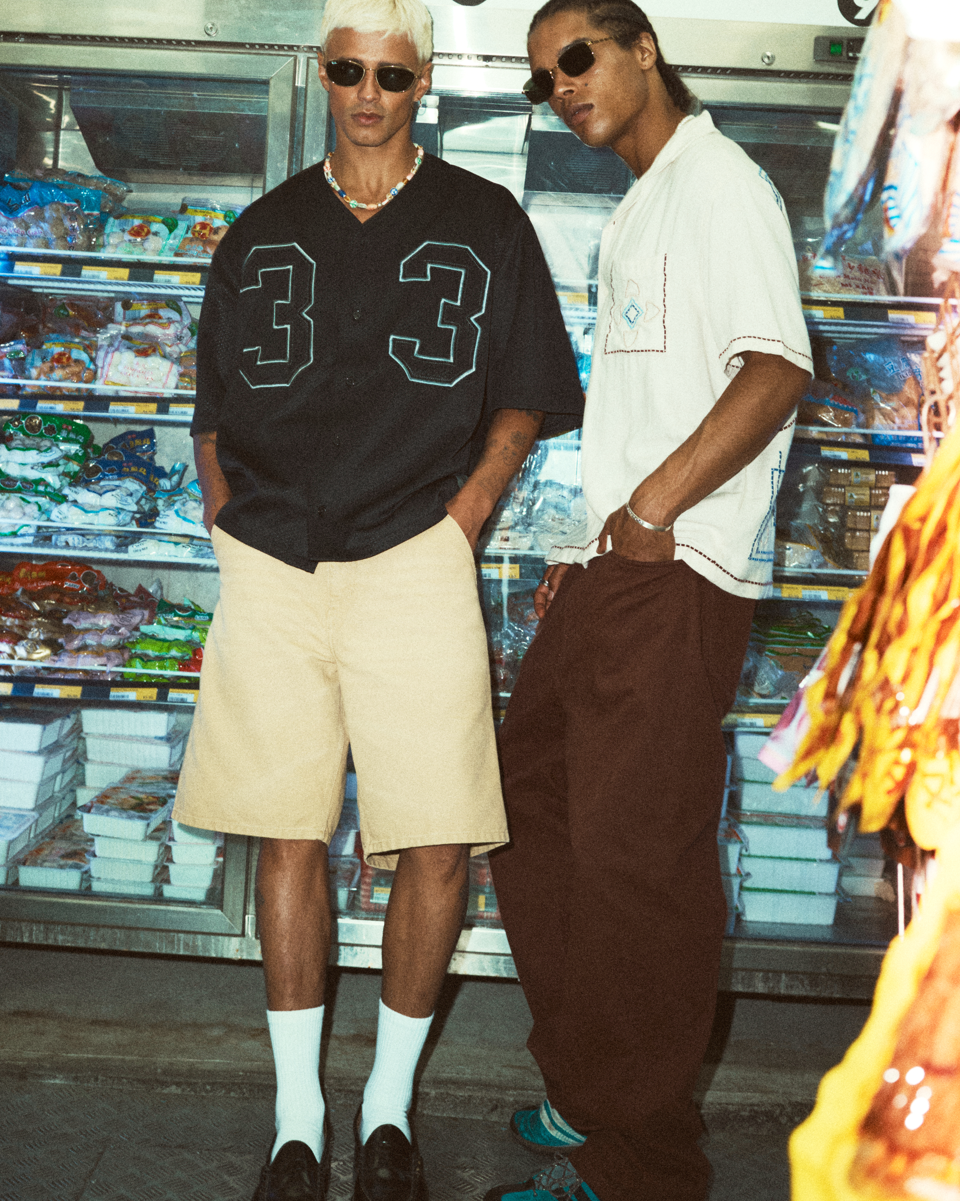 Two young adult men pose indoors in casual fashion, one in a baseball jersey and shorts, the other in an open shirt and pants, cool and casual.