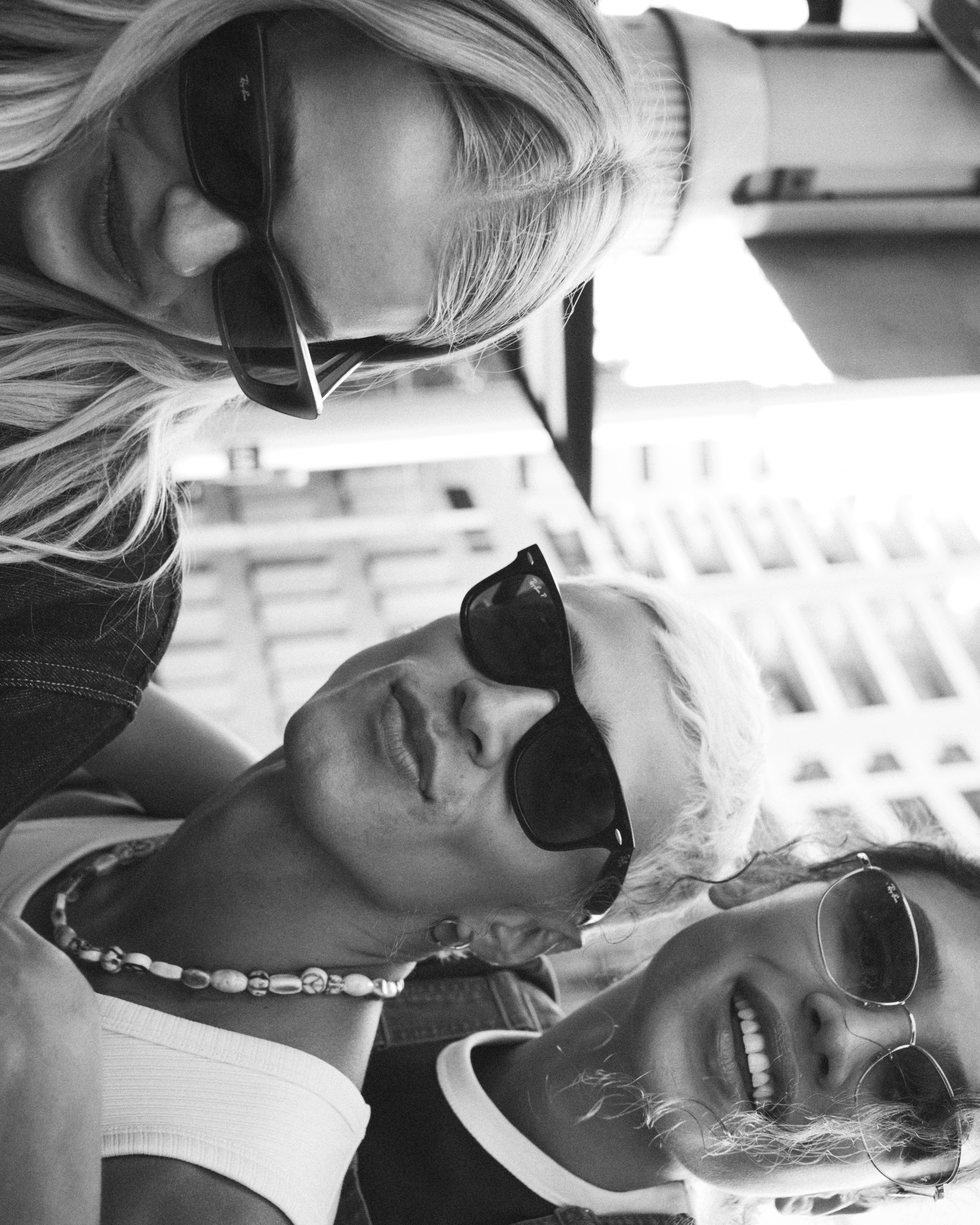 Black and white shot of three young adult women wearing sunglasses and fashionable tops, posing in an urban setting with city buildings.