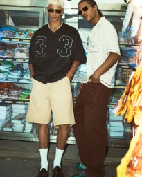 Two male models wearing sunglasses stand in a convenience store aisle in front of refrigerated display cases.