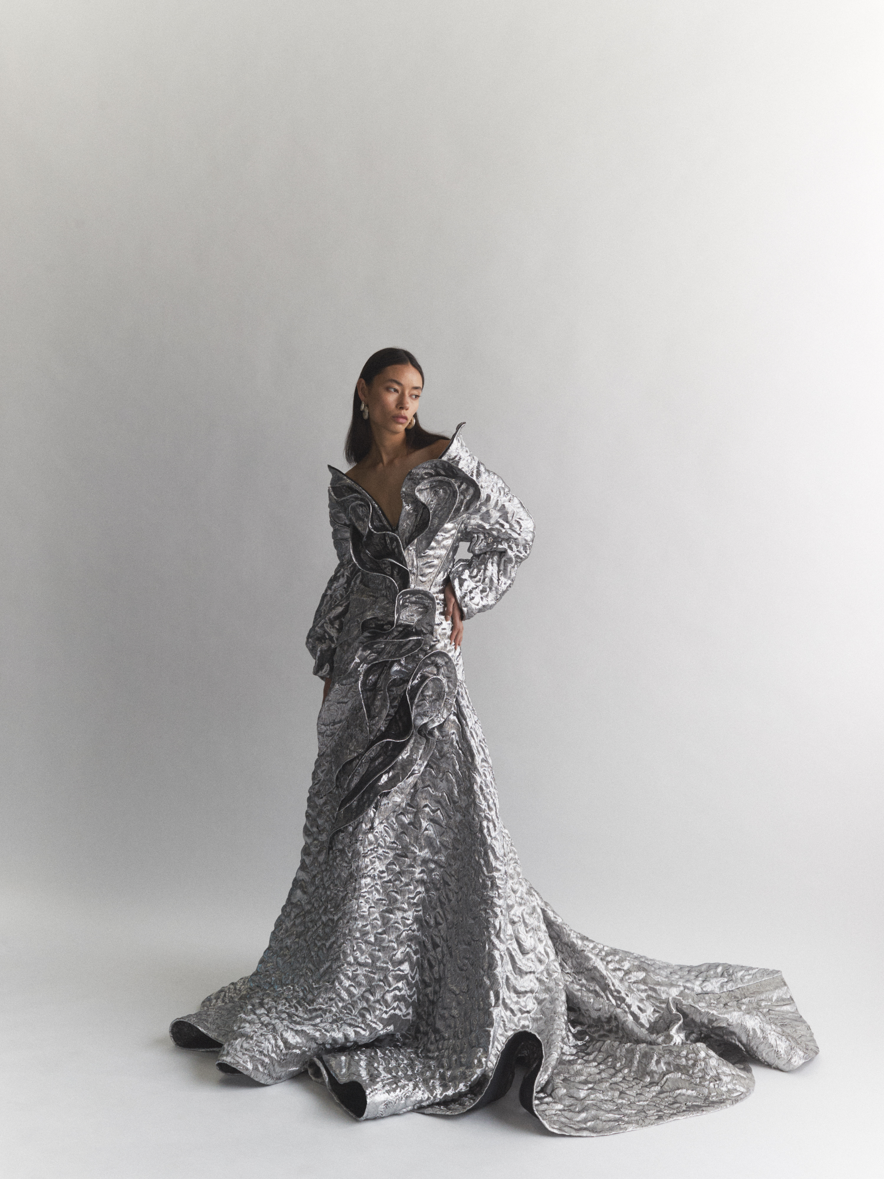 Model wearing a structured metallic silver gown with voluminous ruffled details against a plain white studio background.