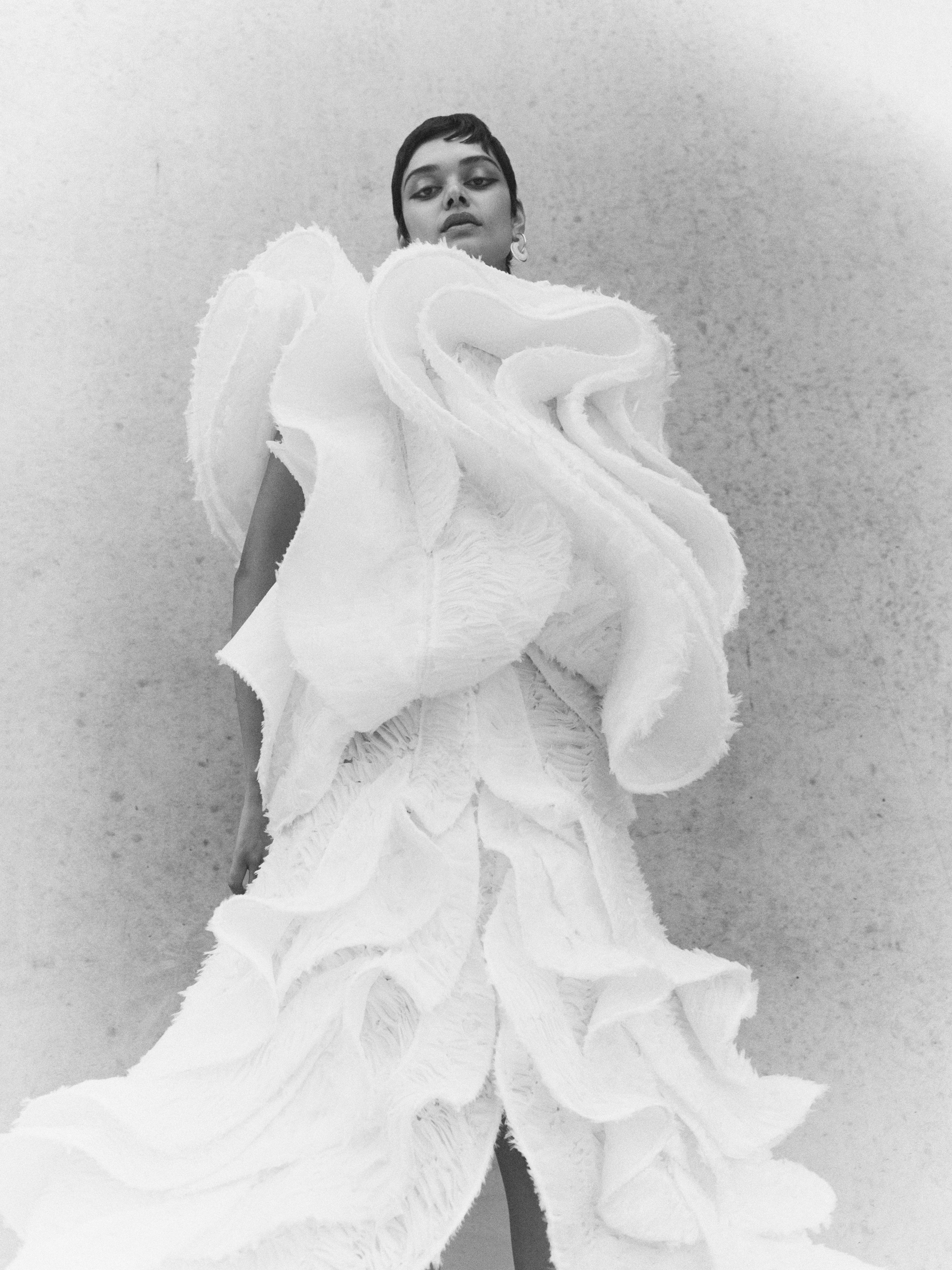 Black and white portrait of a woman wearing an elaborate white ruffled dress in a studio with a textured wall background.