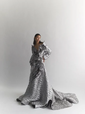 Model wearing a structured metallic silver gown with voluminous ruffled details against a plain white studio background.