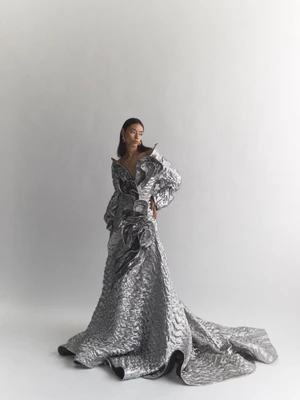 Model wearing a structured metallic silver gown with voluminous ruffled details against a plain white studio background.