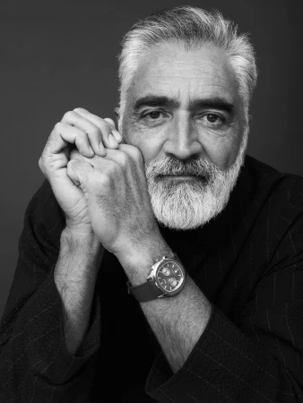 Black and white portrait of an older man with a beard and a watch indoors, with his hands clasped beneath his chin.