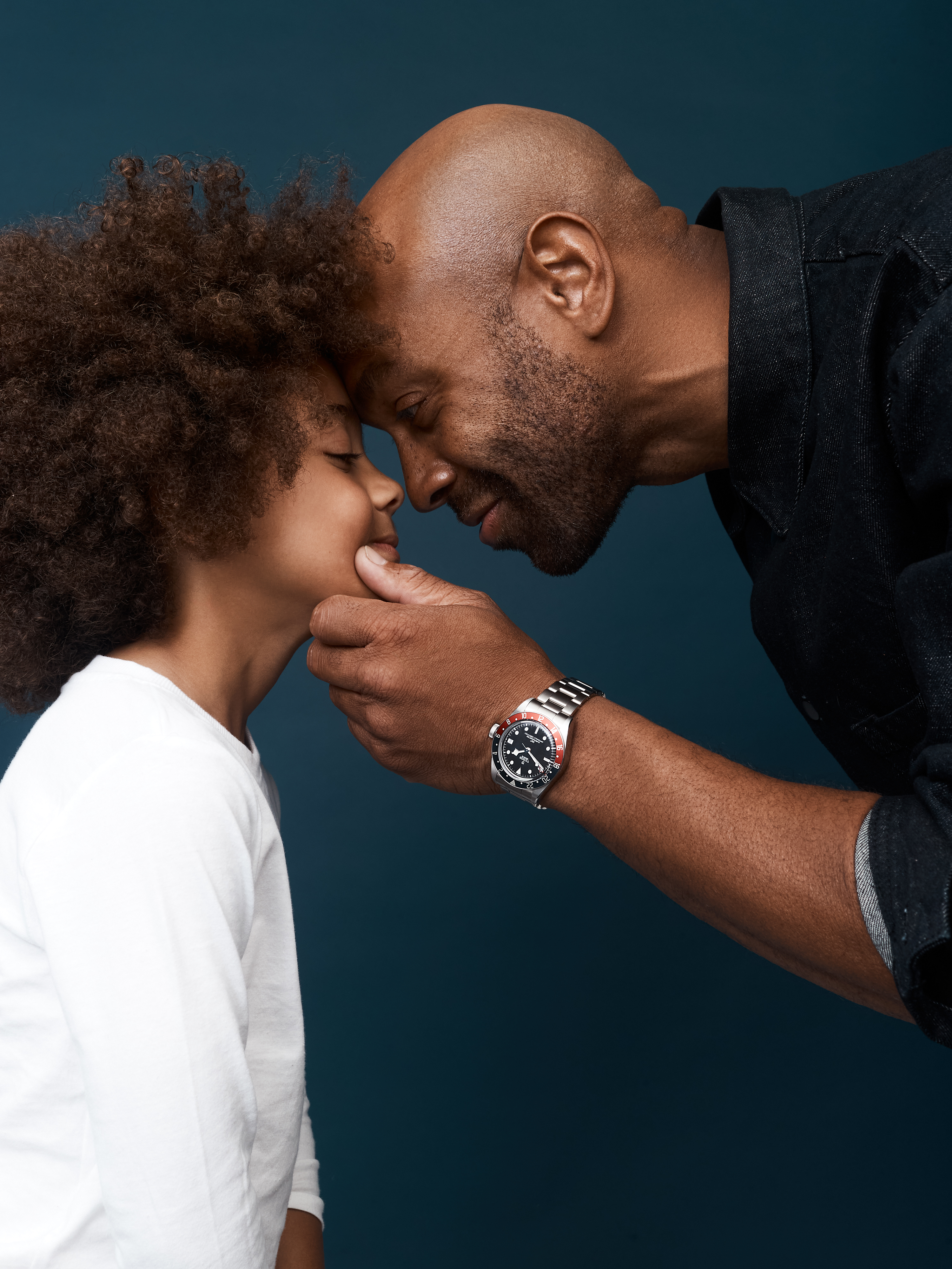 A touching studio portrait shows an African-American father and his young child in a tender moment, eyes closed, against a dark blue backdrop.