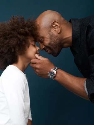 Black father and young child pressing foreheads together in a tender moment against a dark background, photographed by Tim Ashton for Kennedy.