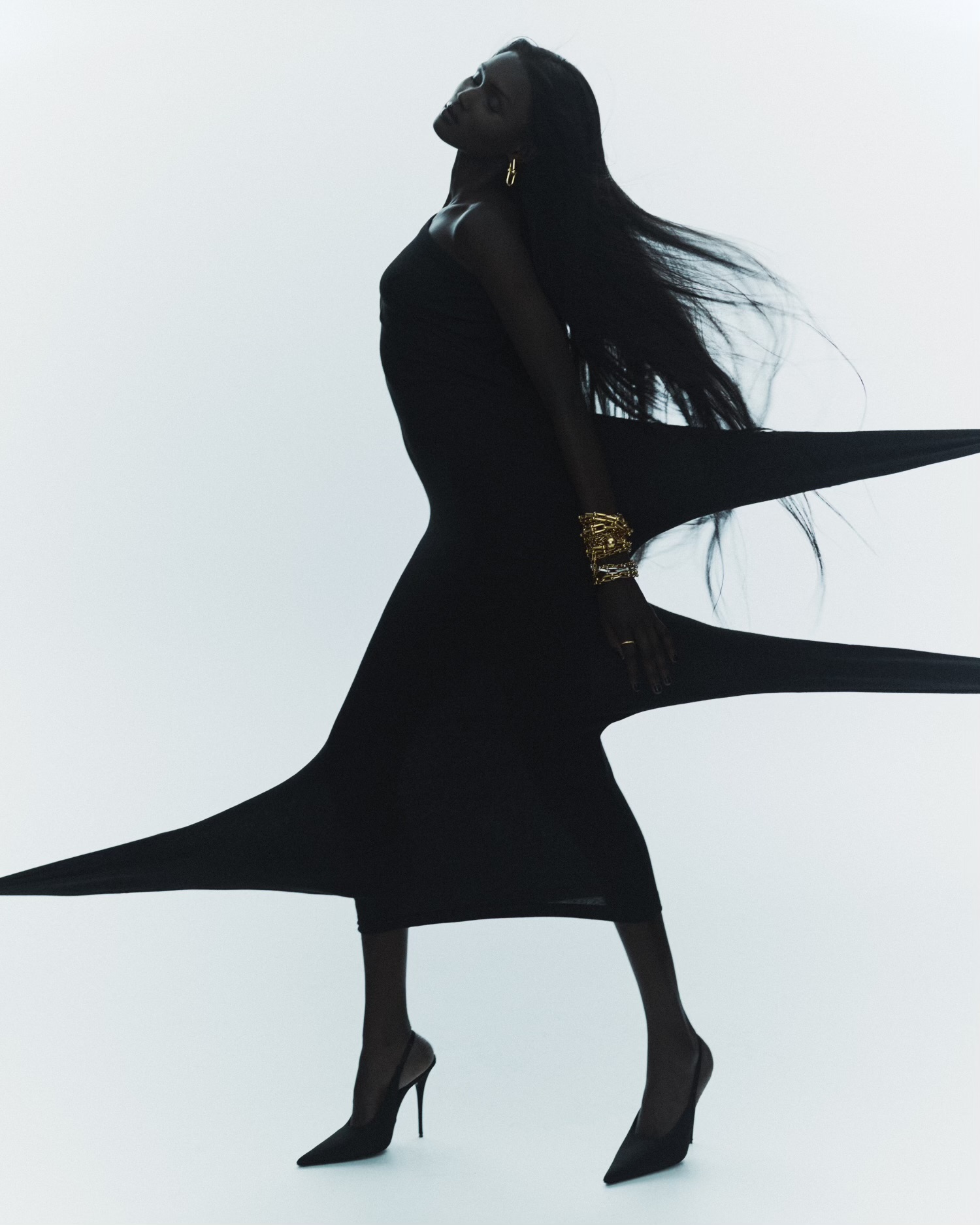 Dark-skinned woman in black dress and heels strikes dramatic pose against a bright white backdrop, accessorized with gold jewelry.
