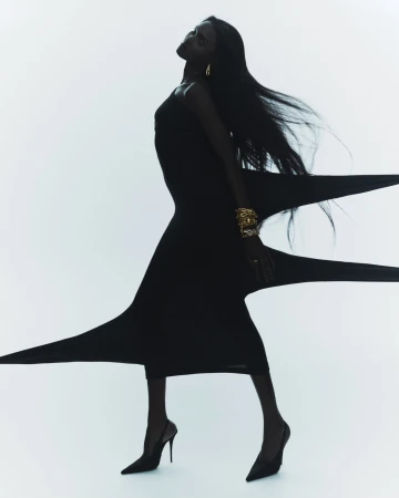 Dark-skinned woman in black dress and heels strikes dramatic pose against a bright white backdrop, accessorized with gold jewelry.