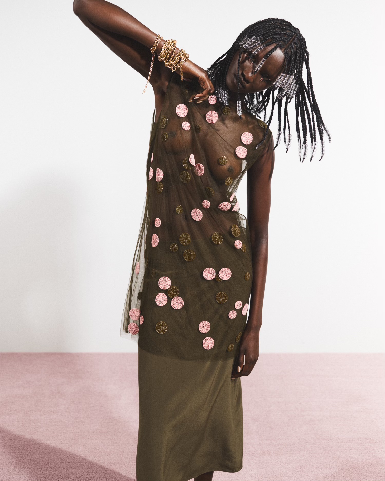 An adult Black woman poses in a sleeveless sheer green dress with pink polka dots against a simple studio backdrop, showcasing fashion and style.
