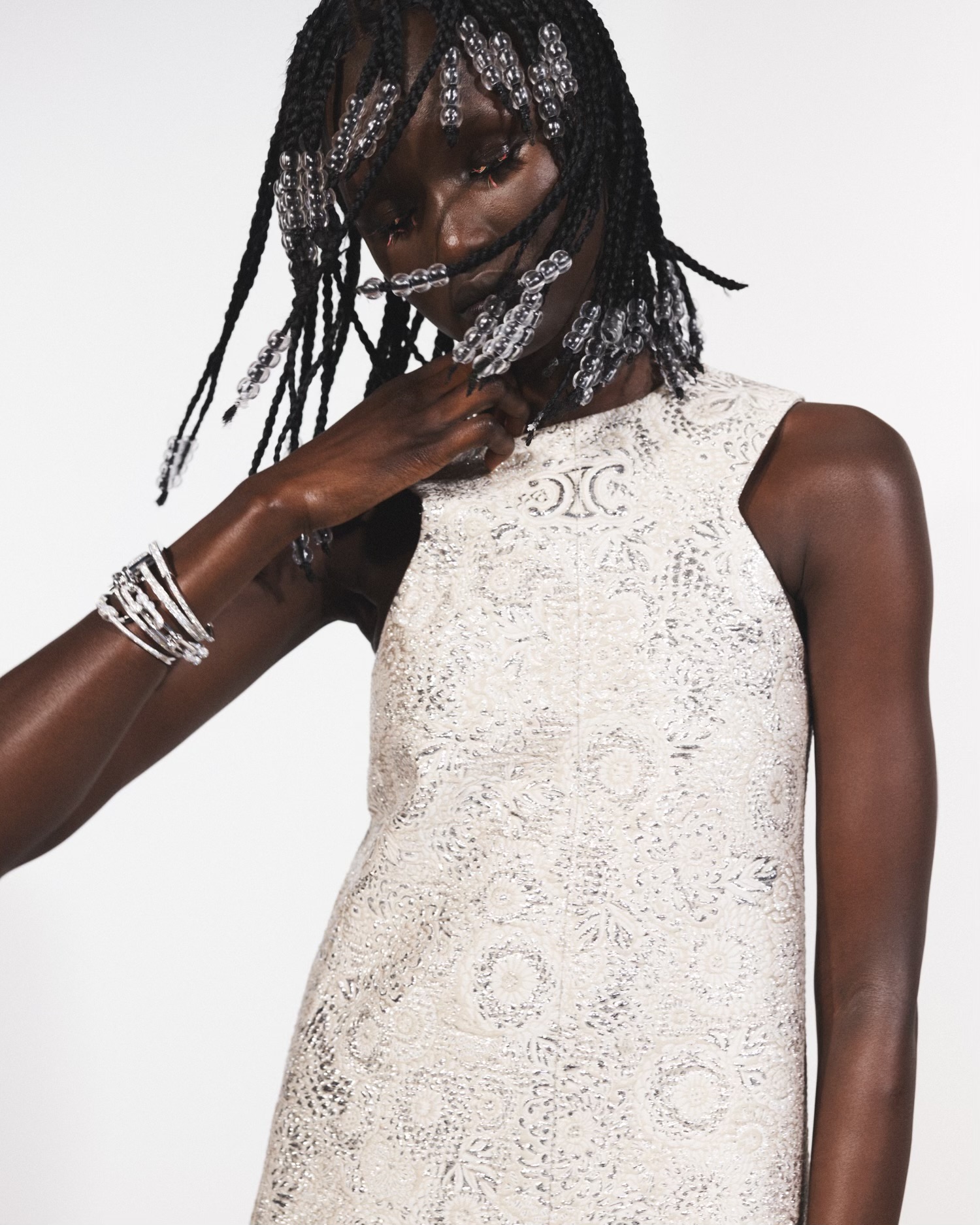 Dark-skinned adult woman in an ivory dress with ornamental braids and silver bracelets posing indoors in front of a plain white backdrop.