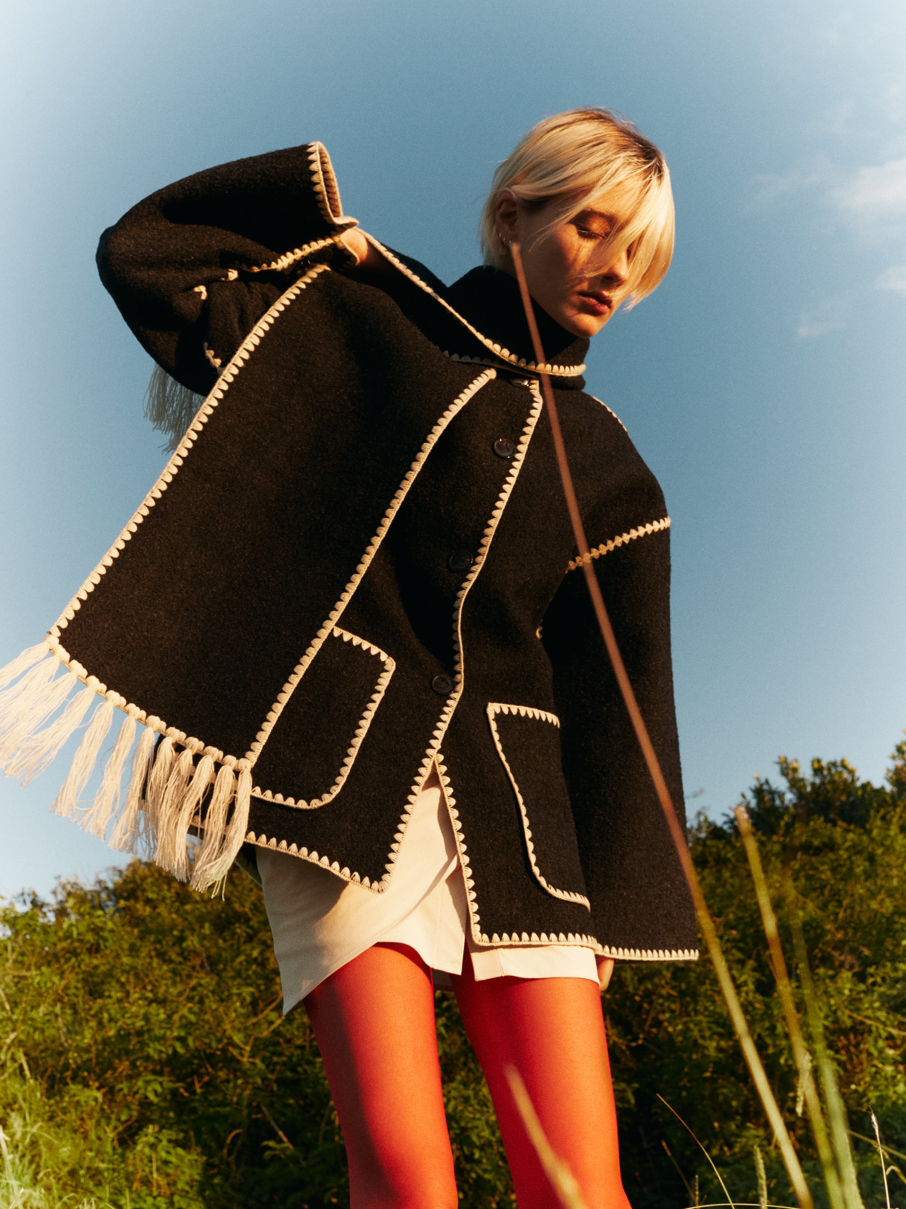 Woman in a black coat and orange stockings poses in a field with a low angle, capturing a fashion-forward look against a natural backdrop.