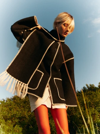 Woman in a black coat and orange stockings poses in a field with a low angle, capturing a fashion-forward look against a natural backdrop.