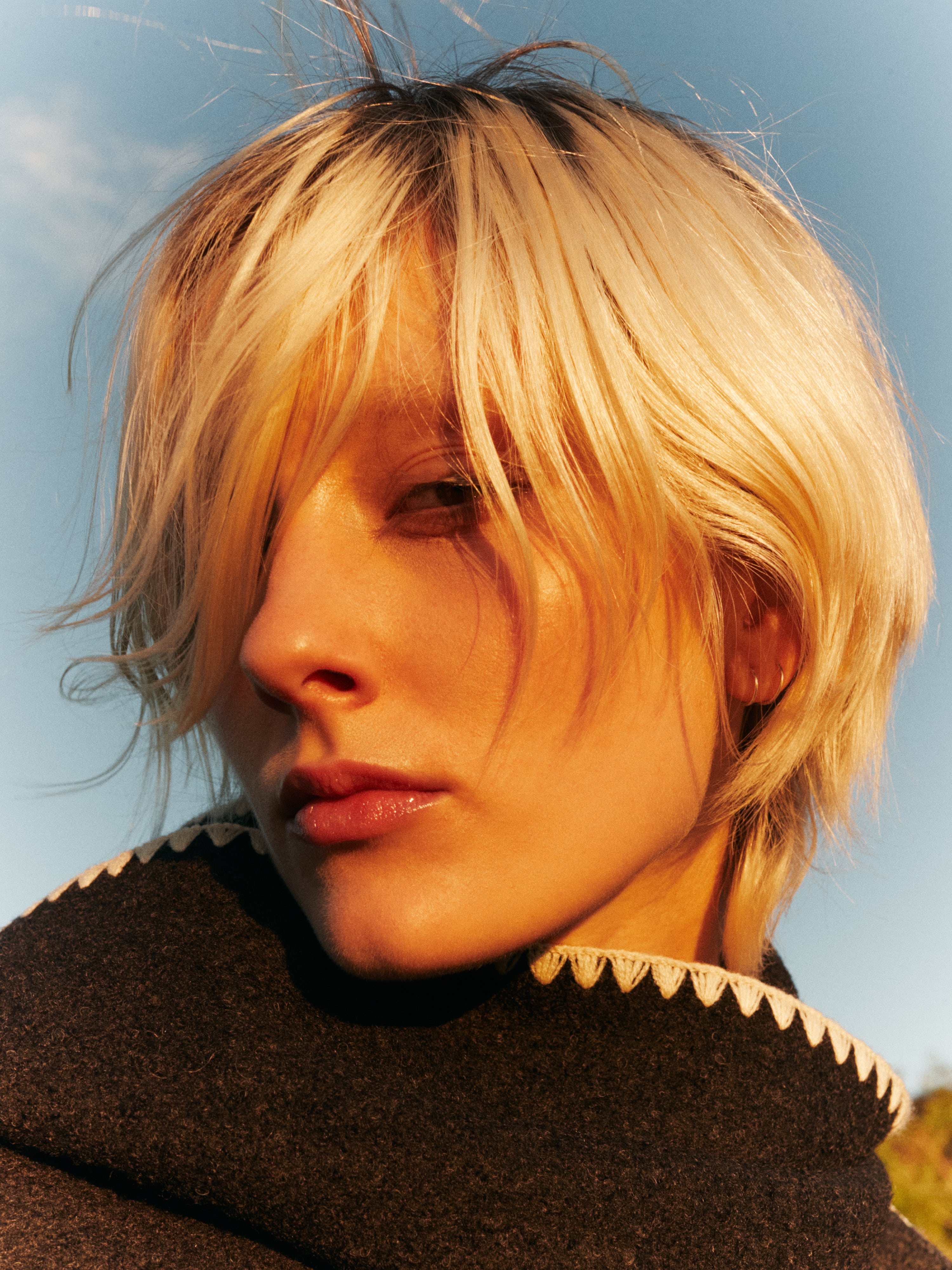 Close-up portrait of a blonde adult female with short hair, fair skin, and a gray coat with a white scalloped trim against a blue sky.