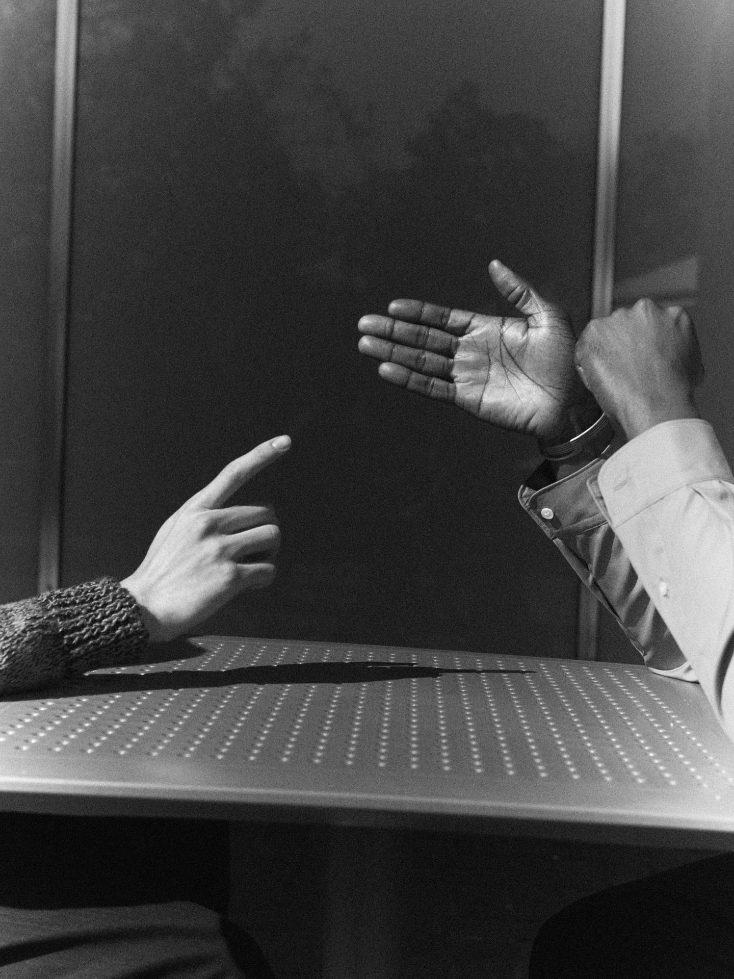High-contrast, black and white image of two hands, one pointing and the other open, meeting over a table against a dark background.