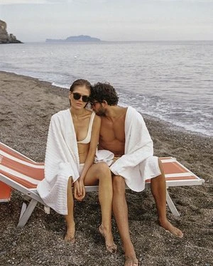 A couple sits on a beach in swimwear and towels on red and white striped lounge chairs with the ocean and an island in the background.