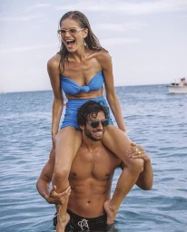 Smiling woman in blue bikini sitting on shoulders of a smiling man in the ocean