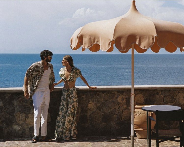 A couple stands on a stone patio overlooking the ocean on a sunny day. The woman wears a floral two-piece outfit. The man wears a patterned shirt.