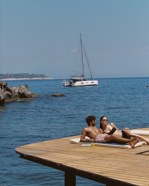 A couple lies on a wooden pier, enjoying the sun with a sailboat in the background, under a clear blue sky, and serene ocean view.