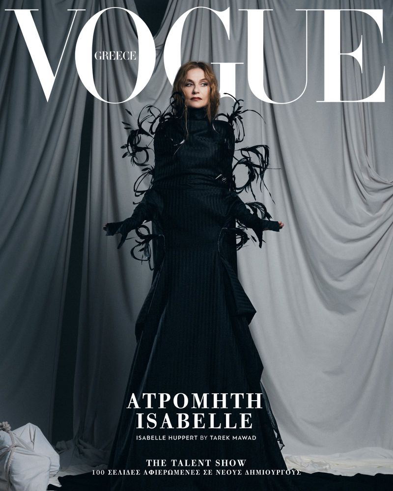 Vogue Greece cover featuring Isabelle Huppert in a black gown with feathered accents, set against a draped, light grey fabric backdrop.