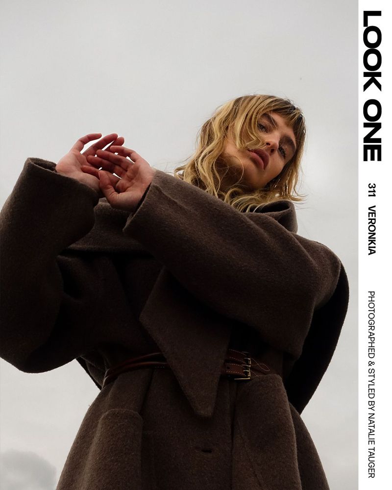 Fashion editorial shot of a female model wearing a brown wool coat, posing with hands near her face against a muted sky backdrop.