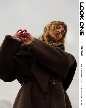 Model Veronika Vilim in a brown belted coat shot from a low angle against an overcast sky