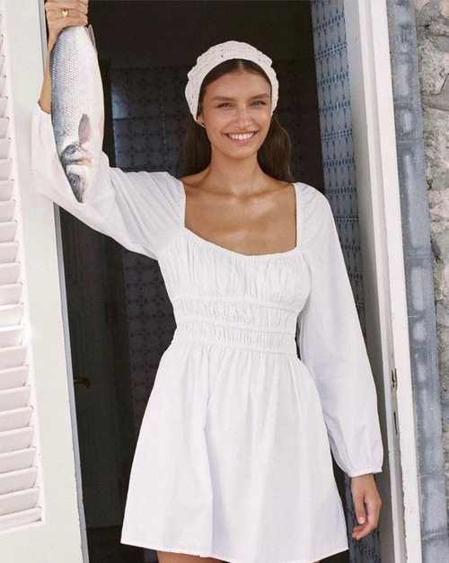Smiling woman in a white dress holds a fresh fish in a doorway, bathed in natural light. The setting creates a summery fashion vibe.