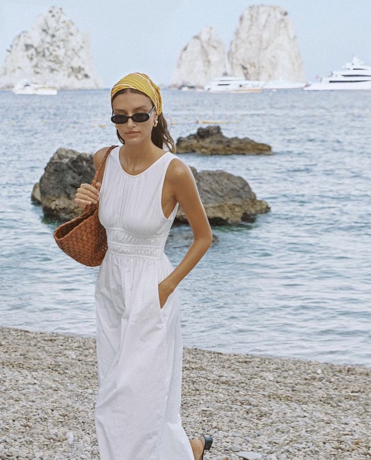 A woman in a white dress and yellow headscarf poses on a rocky beach with islands and yachts visible in the background on a sunny day.